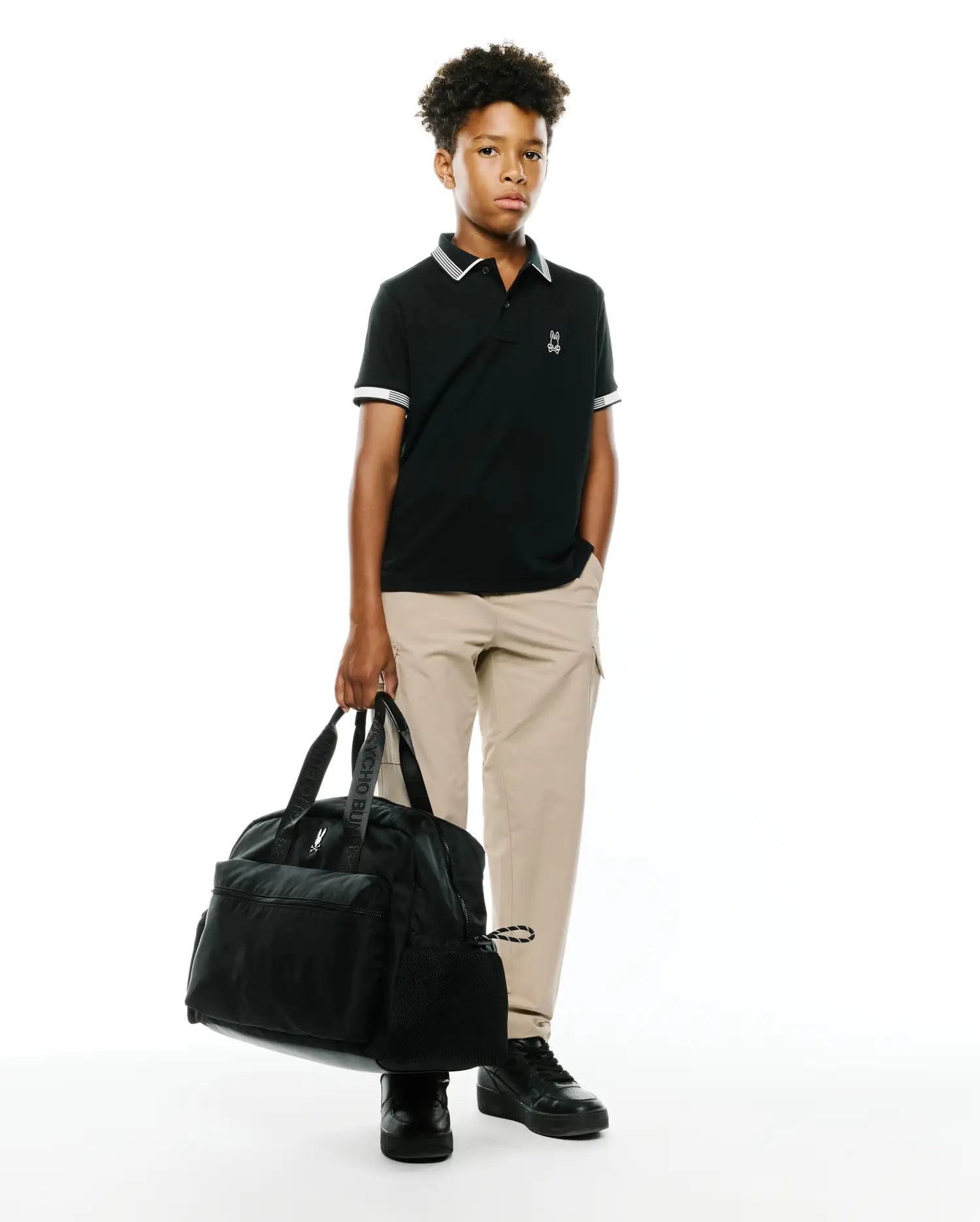 Kids Timmy Pique Polo in classic black pique with white striped collar and cuffs, featuring a small embroidered white Psycho Bunny skull logo on the left chest, styled with khaki pants.