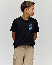 A boy with short brown hair wears the Psycho Bunny KIDS PUERTO RICO TEE (KT0200133), a black Pima cotton shirt with a vibrant palm tree graphic, and beige cargo shorts. He stands with hands behind his back, looking left against a plain light background.