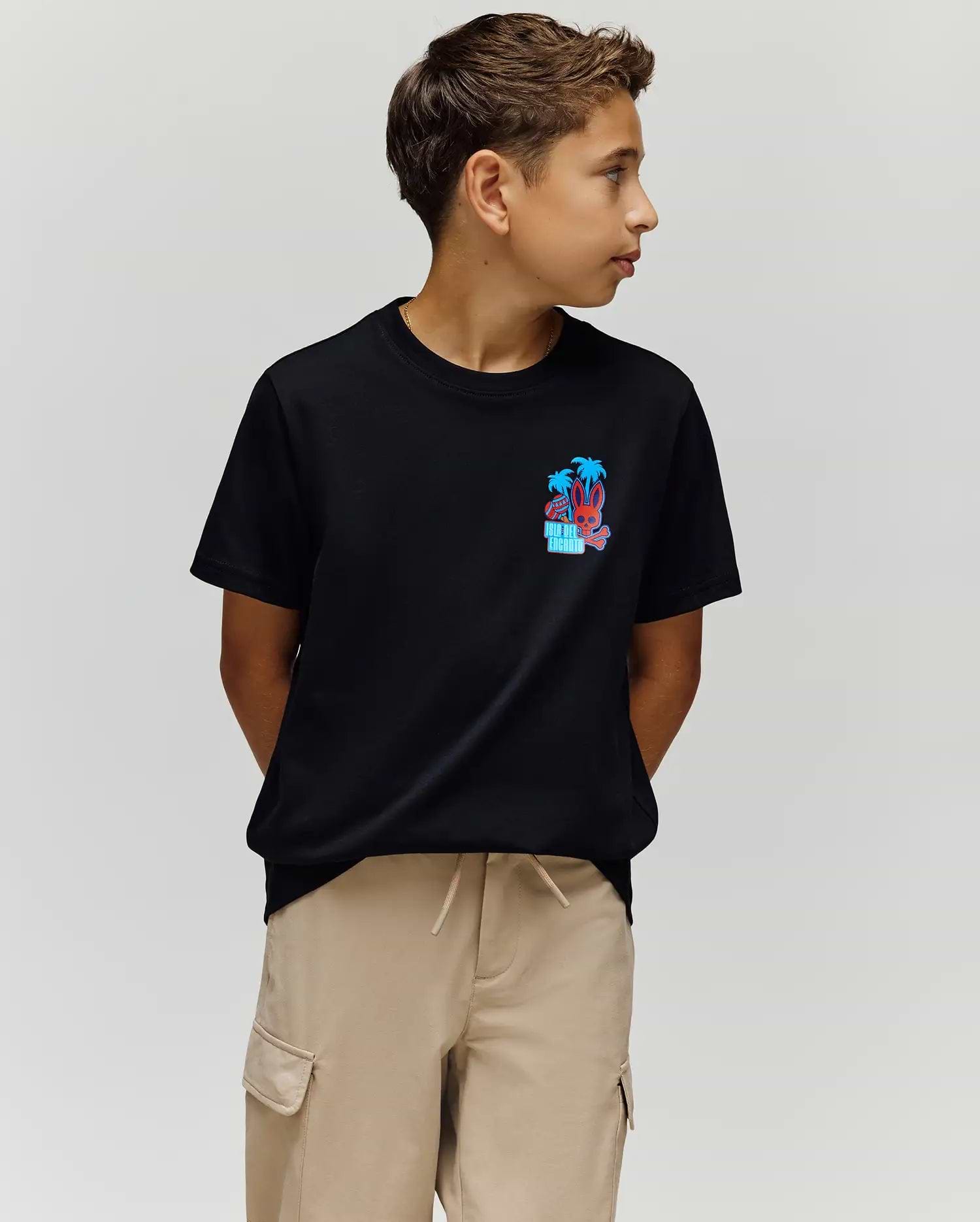 A boy with short brown hair wears the Psycho Bunny KIDS PUERTO RICO TEE (KT0200133), a black Pima cotton shirt with a vibrant palm tree graphic, and beige cargo shorts. He stands with hands behind his back, looking left against a plain light background.