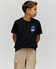 A boy with short brown hair wears the Psycho Bunny KIDS PUERTO RICO TEE (KT0200133), a black Pima cotton shirt with a vibrant palm tree graphic, and beige cargo shorts. He stands with hands behind his back, looking left against a plain light background.