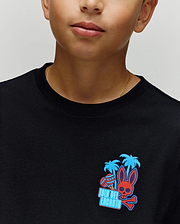 A child wears the Psycho Bunny KIDS PUERTO RICO TEE (KT0200133), a soft black Pima cotton shirt featuring vibrant blue palms, a red skull bunny graphic, and "Isla Del Encanto" text—embodying Puerto Rico’s lively spirit.