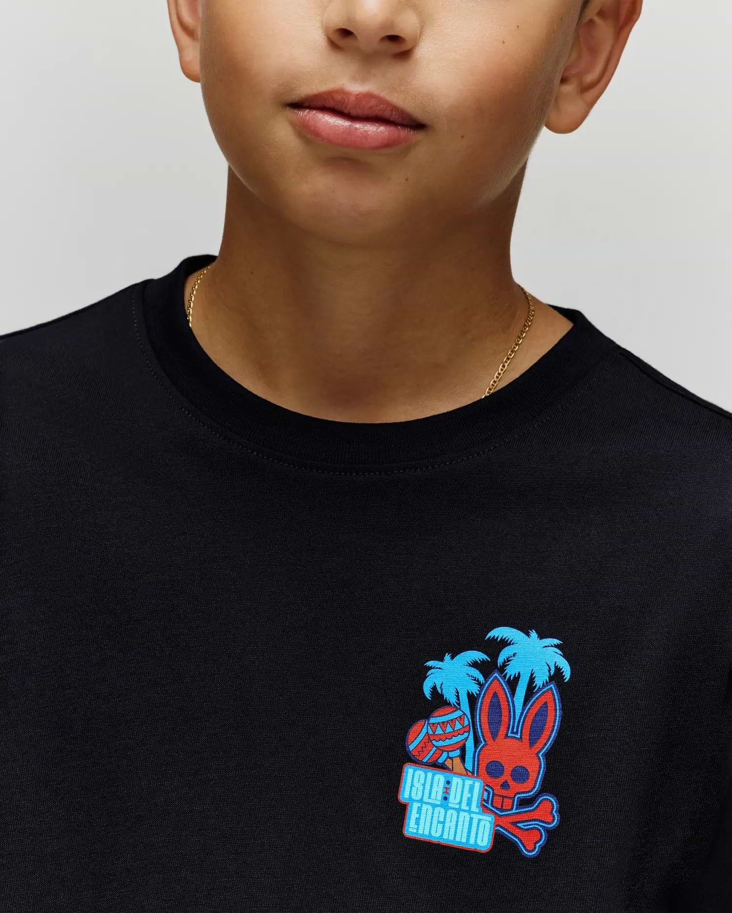 A child wears the Psycho Bunny KIDS PUERTO RICO TEE (KT0200133), a soft black Pima cotton shirt featuring vibrant blue palms, a red skull bunny graphic, and "Isla Del Encanto" text—embodying Puerto Rico’s lively spirit.