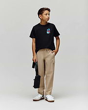 A young boy models the Psycho Bunny KIDS PUERTO RICO TEE (KT0200133), a black Pima cotton t-shirt with a colorful bunny graphic, paired with beige cargo pants, white sneakers, and a black bag, looking to his right against a plain backdrop.