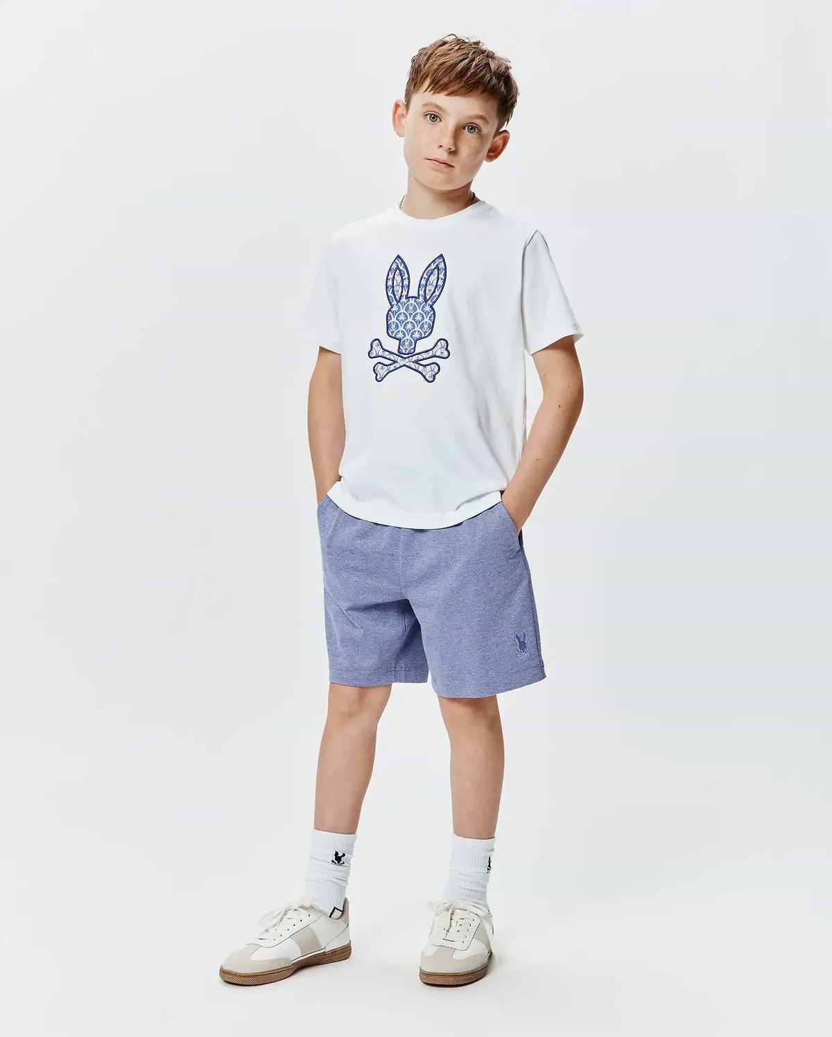 Kids Brad Graphic Tee, a white jersey tee with a large ornate blue Psycho Bunny skull graphic, paired with heather blue relaxed knit shorts featuring a small embroidered bunny logo.