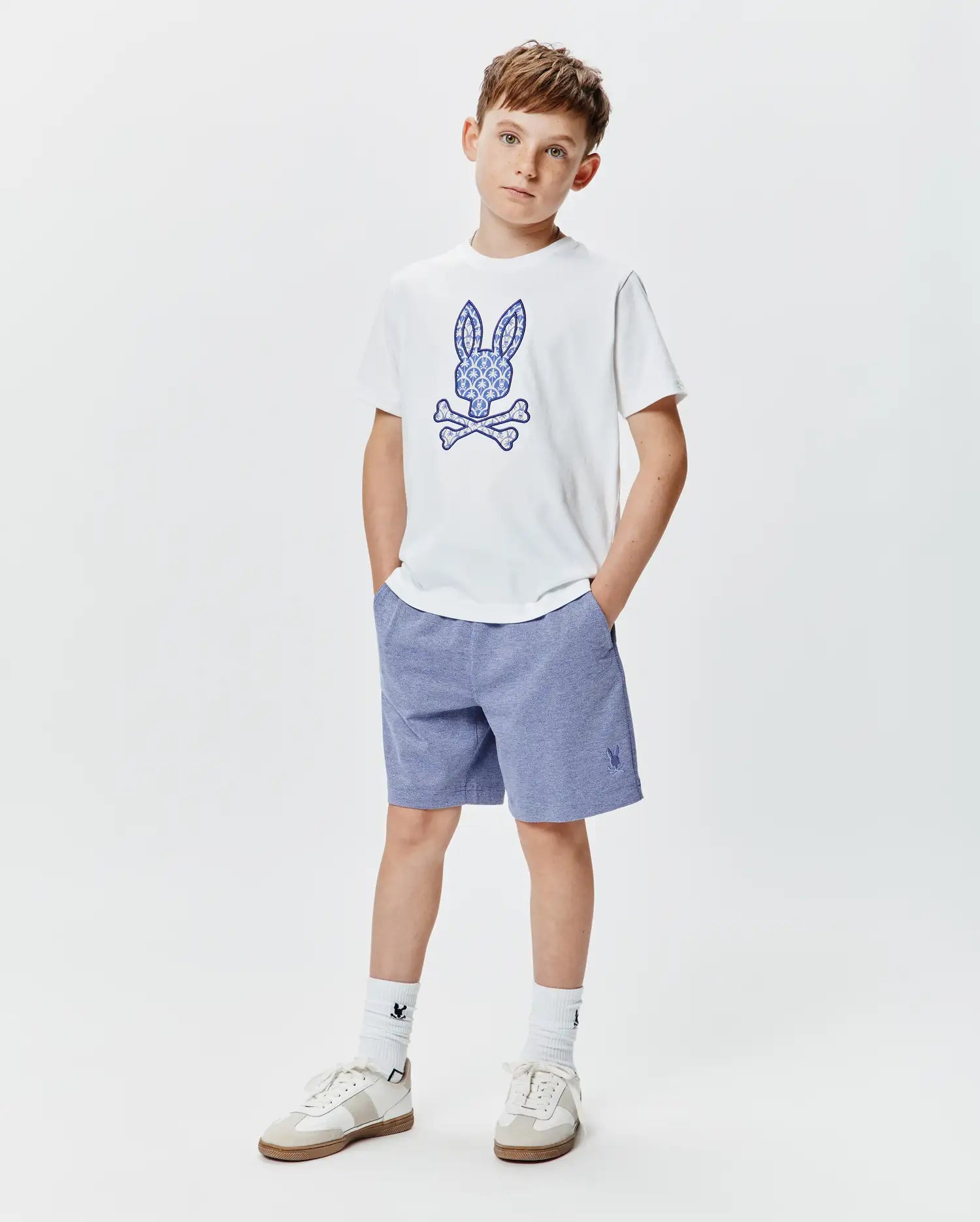Kids Brad Graphic Tee, a white jersey tee with a large ornate blue Psycho Bunny skull graphic, paired with heather blue relaxed knit shorts featuring a small embroidered bunny logo.