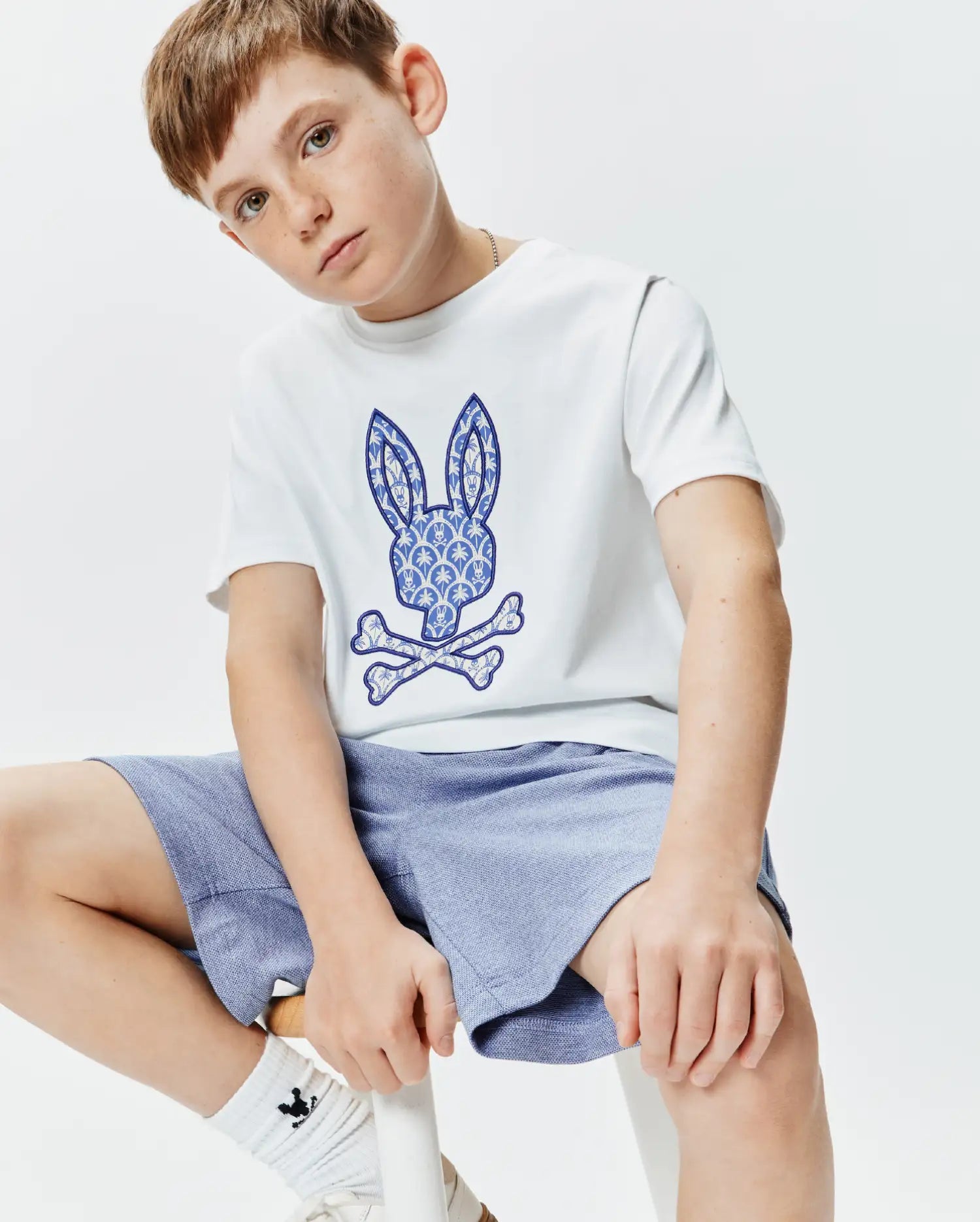 White Kids Brad Graphic Tee, classic fit jersey, with a large blue and white palm tree patterned Psycho Bunny skull and crossbones logo on the center chest.