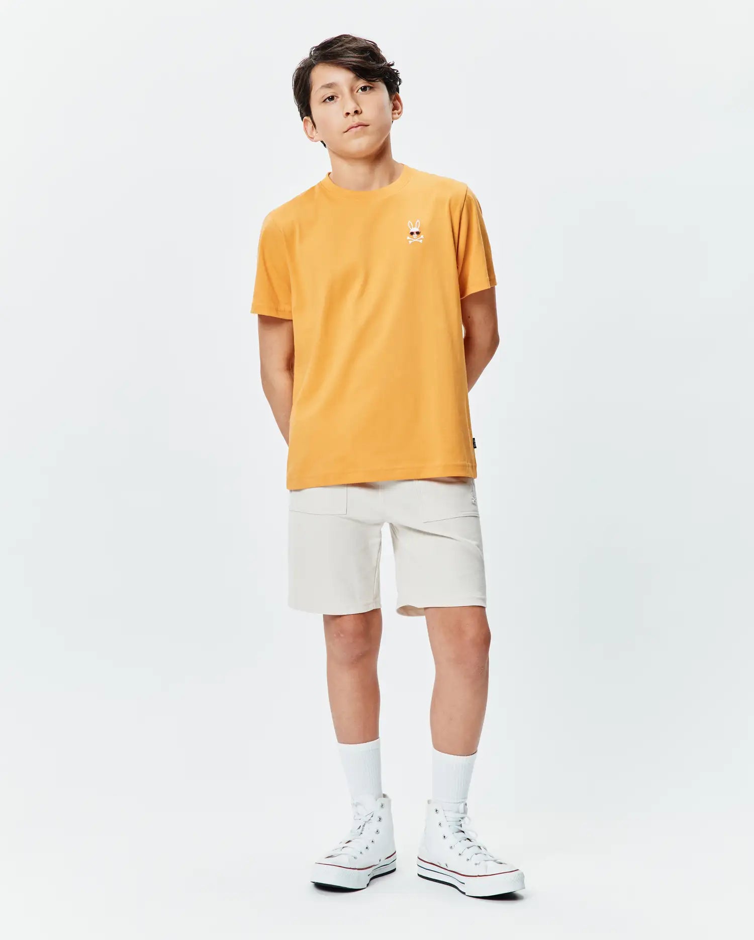 Orange Kids Sunny Chest Graphic Tee in classic fit jersey, featuring a small embroidered Psycho Bunny skull logo on the left chest, paired with off-white woven shorts.