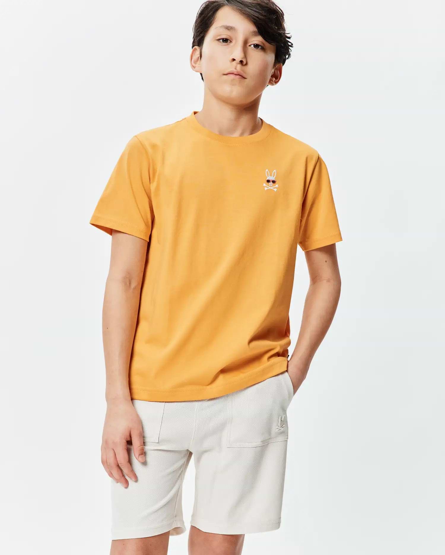 Kids Sunny Chest Graphic Tee, a classic fit sunny orange jersey t-shirt featuring a small embroidered Psycho Bunny skull with sunglasses and crossbones on the left chest.