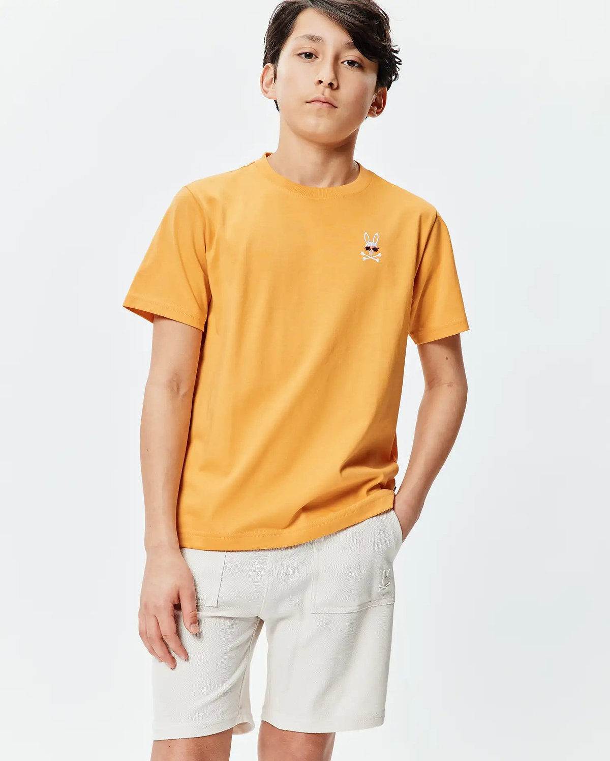 Kids Sunny Chest Graphic Tee, a classic fit sunny orange jersey t-shirt featuring a small embroidered Psycho Bunny skull with sunglasses and crossbones on the left chest.