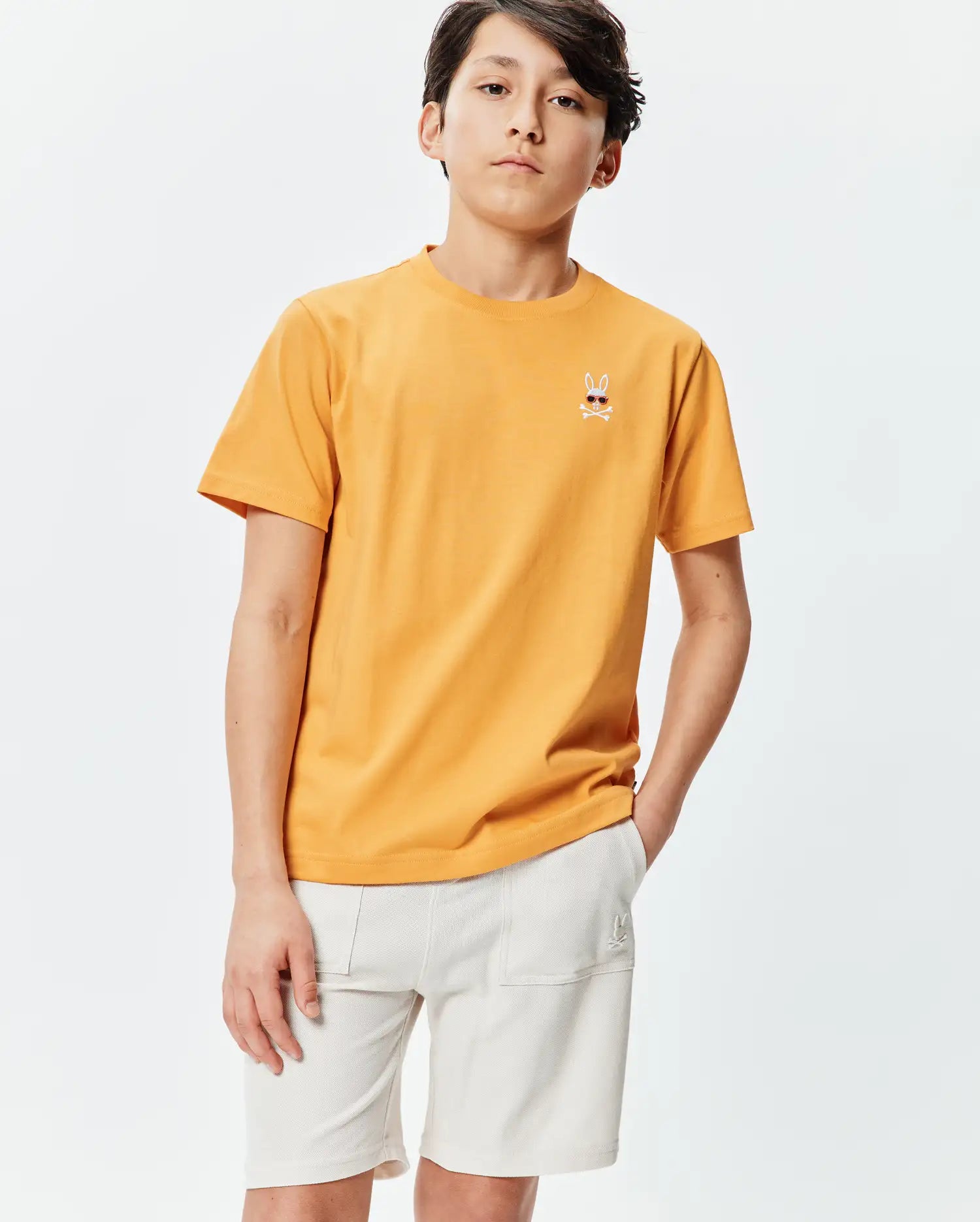 Kids Sunny Chest Graphic Tee, a classic fit sunny orange jersey t-shirt featuring a small embroidered Psycho Bunny skull with sunglasses and crossbones on the left chest.