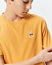 Kids Sunny Chest Graphic Tee in mustard yellow jersey knit, featuring a small embroidered Psycho Bunny skull logo with red sunglasses and white crossbones on the left chest, classic fit.