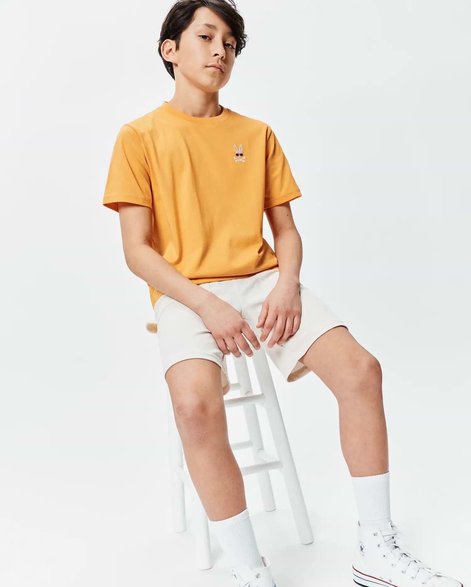 Kids Sunny Chest Graphic Tee in sunny yellow, featuring a classic fit and smooth jersey fabric. It has a small embroidered Psycho Bunny skull logo with red sunglasses on the left chest, paired with cream relaxed-fit shorts.