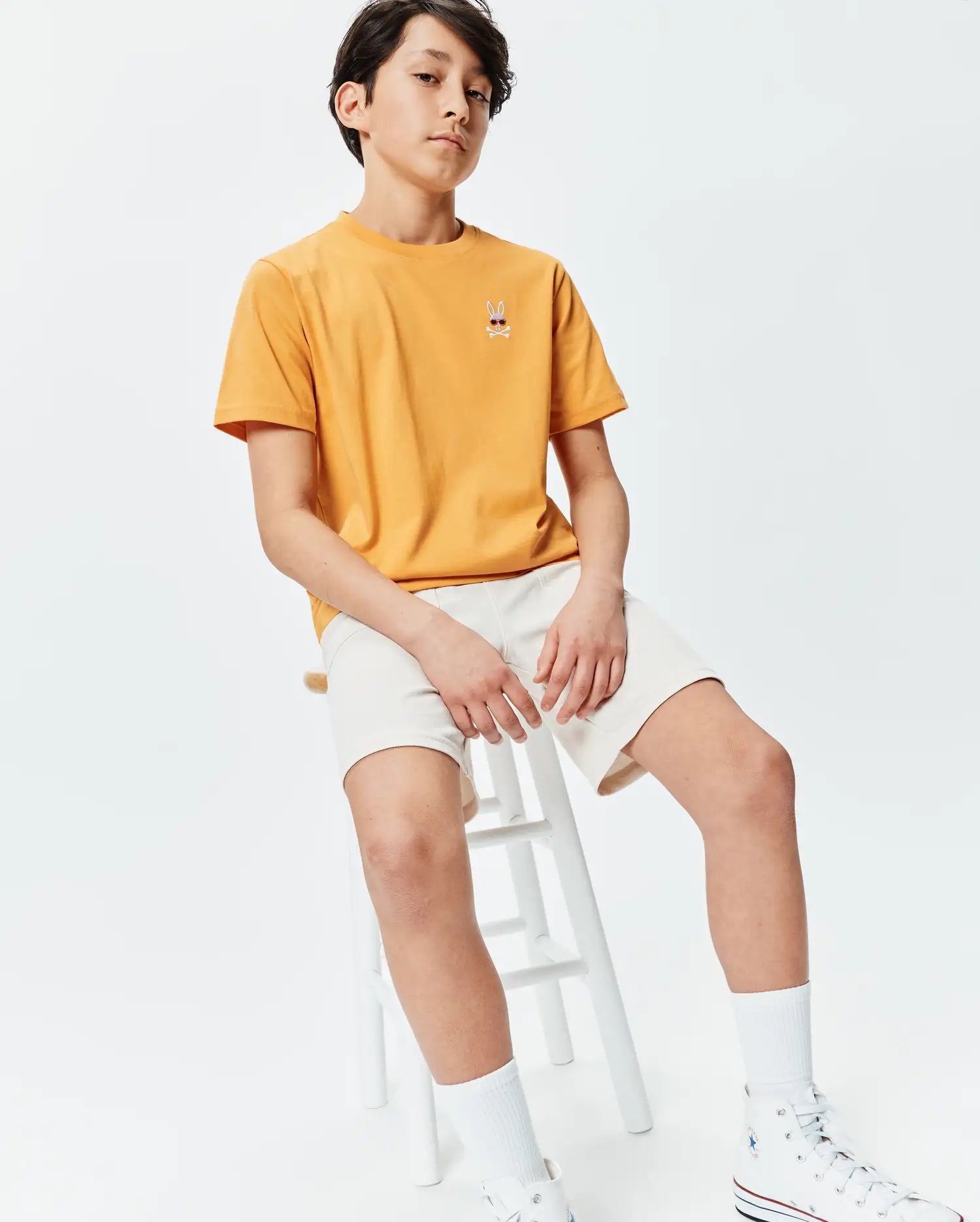 Kids Sunny Chest Graphic Tee in sunny yellow, featuring a classic fit and smooth jersey fabric. It has a small embroidered Psycho Bunny skull logo with red sunglasses on the left chest, paired with cream relaxed-fit shorts.