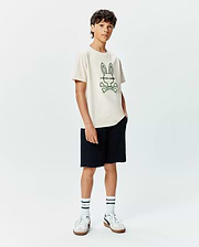 Kids Craig Graphic Tee in cream, featuring a large embroidered green Psycho Bunny skull and crossbones logo with text on the center chest, paired with black shorts. Classic fit jersey knit.