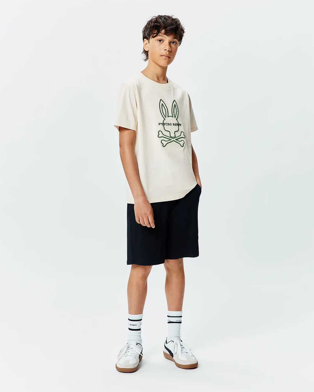 Kids Craig Graphic Tee in cream, featuring a large embroidered green Psycho Bunny skull and crossbones logo with text on the center chest, paired with black shorts. Classic fit jersey knit.