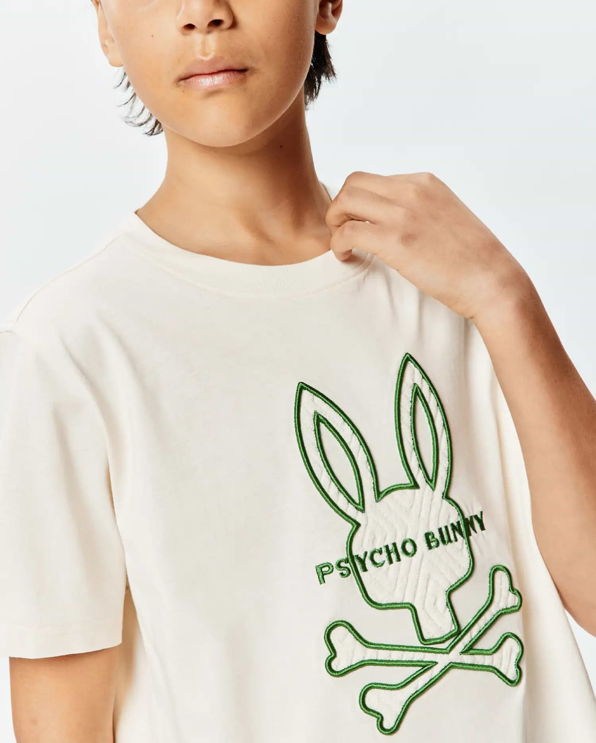 Kids Craig Graphic Tee, an off-white jersey t-shirt with a large, textured cream and green embroidered Psycho Bunny skull logo on the chest, featuring green outline and green text.