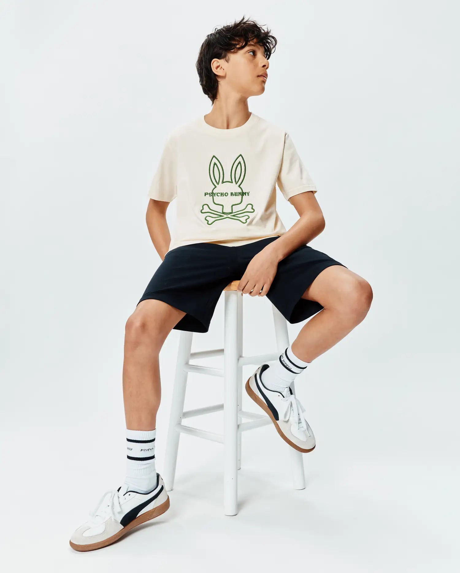 Kids Craig Graphic Tee in cream jersey, featuring a large central green Psycho Bunny skull and crossbones graphic print, paired with classic fit black shorts.