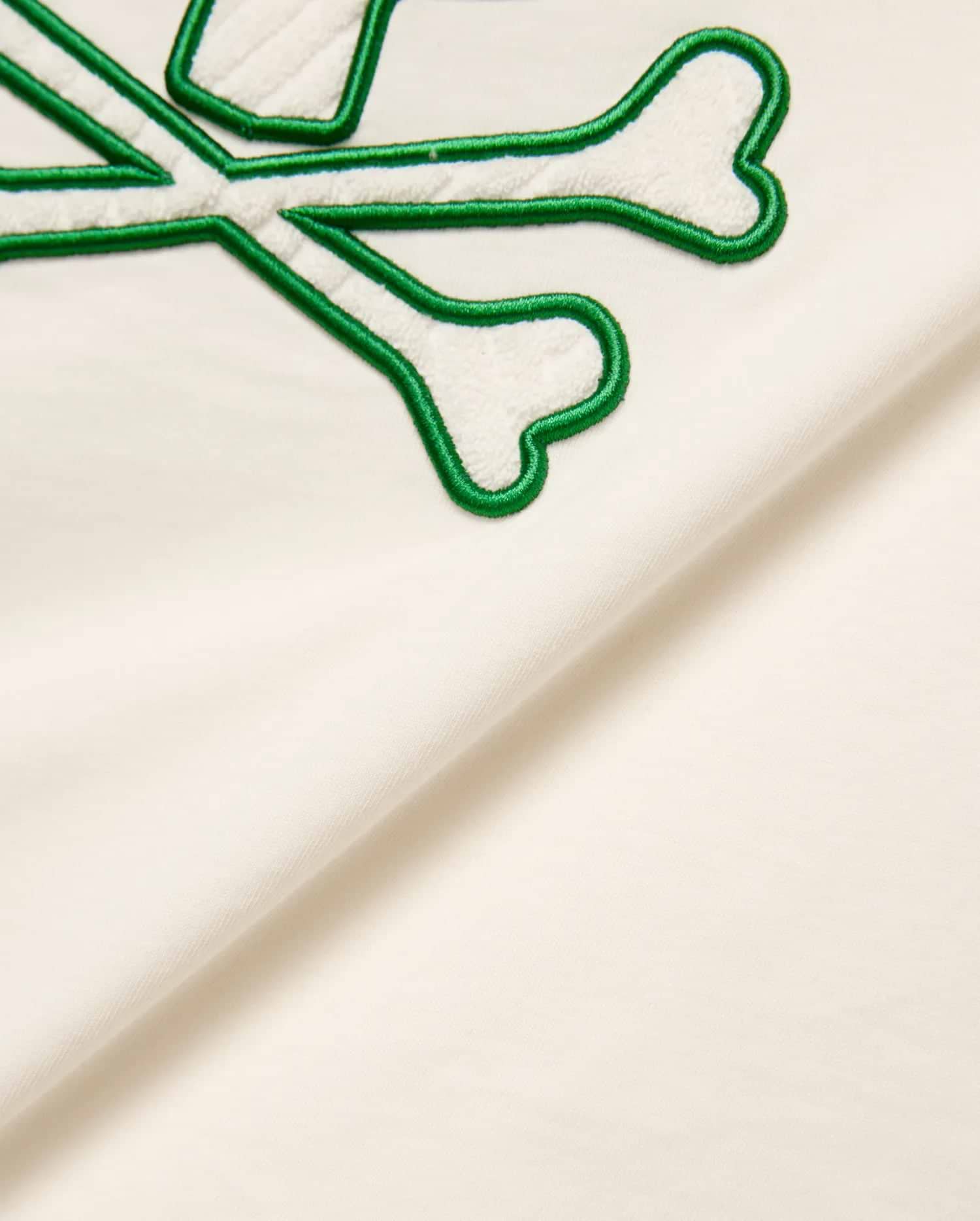 Cream Kids Craig Graphic Tee featuring a prominent green-outlined, white terry cloth embroidered Psycho Bunny skull and crossbones logo on soft jersey fabric.