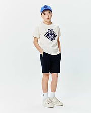 Kids Tobias Graphic Tee in cream jersey, featuring a large central dark blue Psycho Bunny skull and crossbones logo within a diamond, paired with navy shorts and a blue cap.