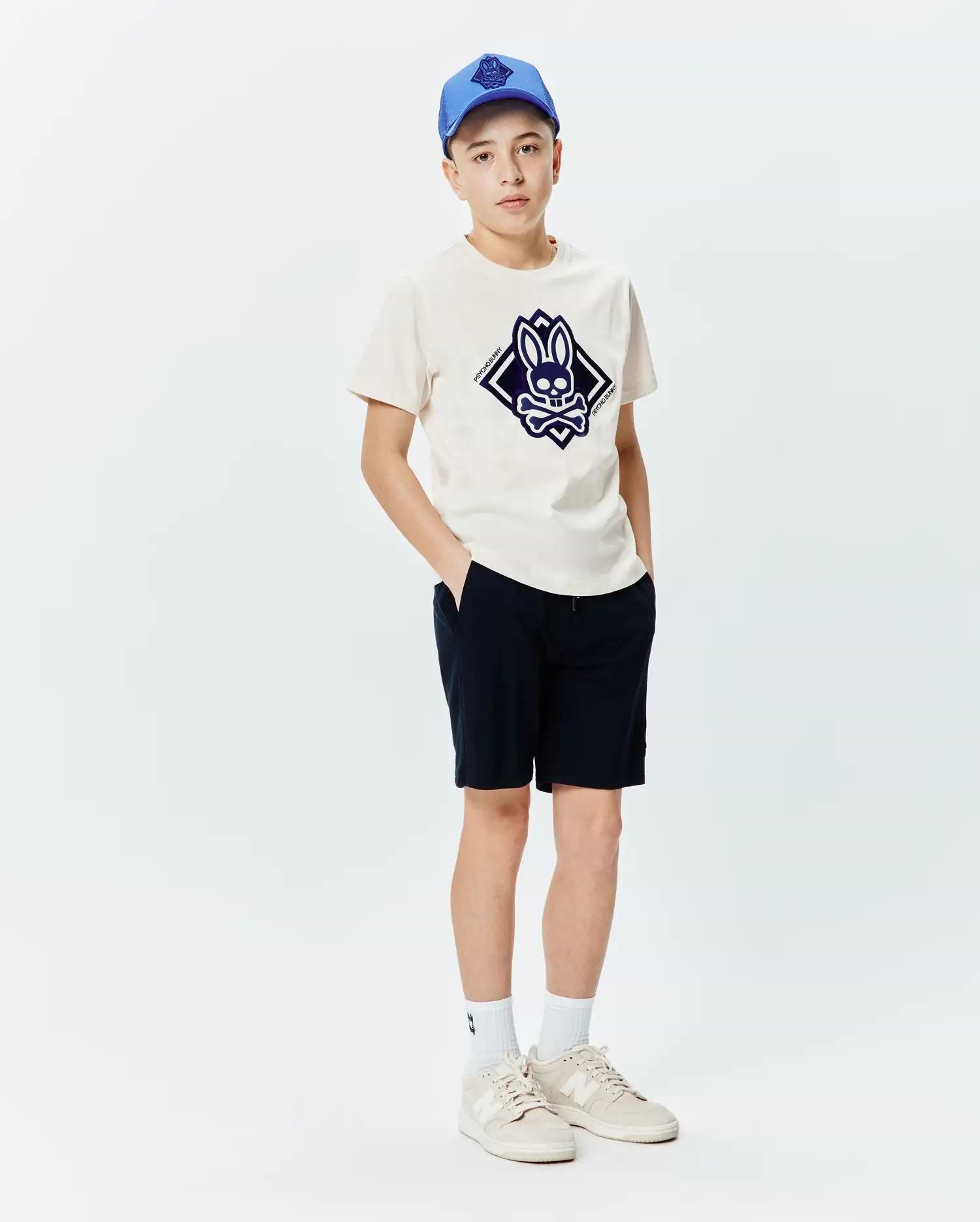 Kids Tobias Graphic Tee in cream jersey, featuring a large central dark blue Psycho Bunny skull and crossbones logo within a diamond, paired with navy shorts and a blue cap.