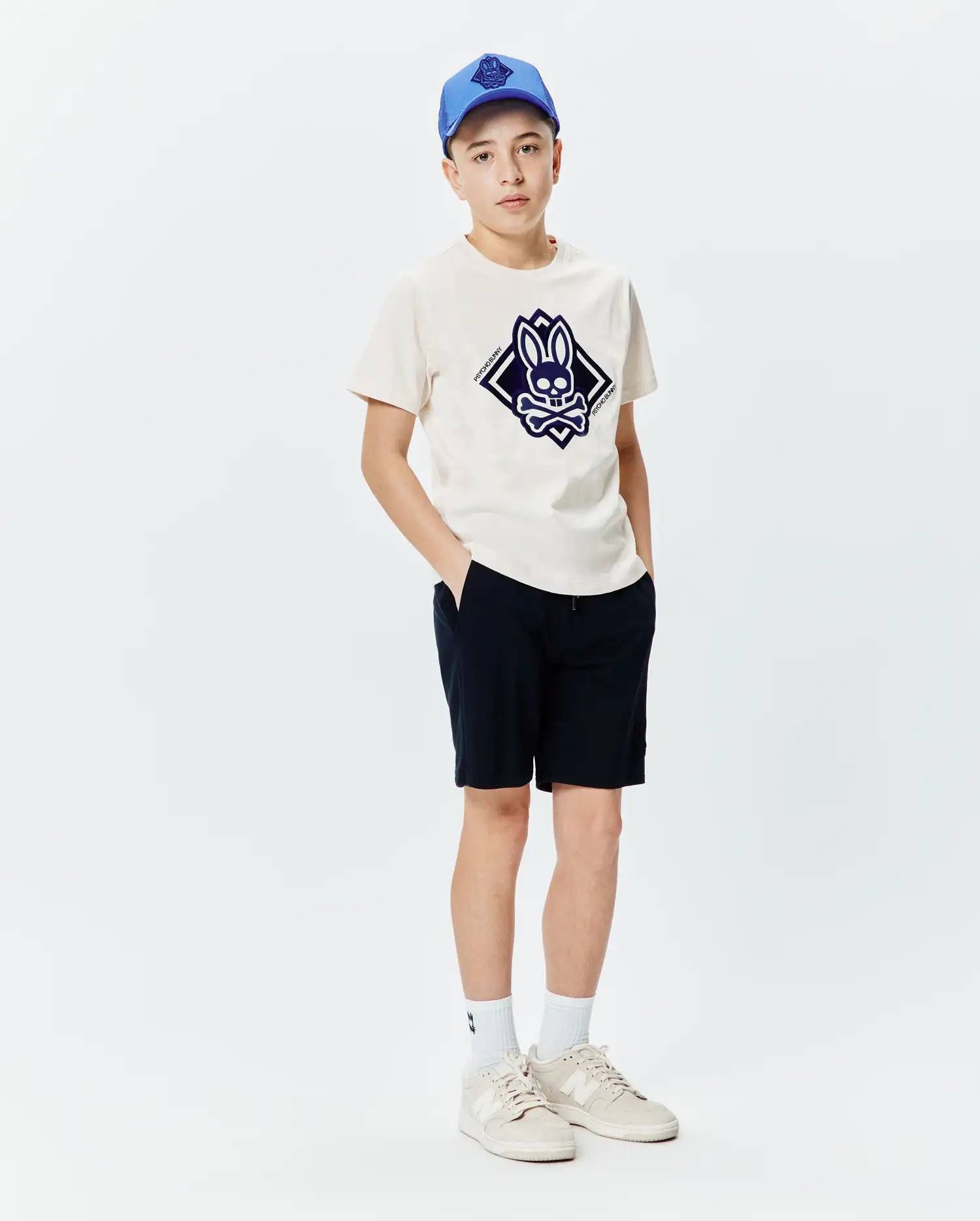 Kids Tobias Graphic Tee in cream jersey, featuring a large central dark blue Psycho Bunny skull and crossbones logo within a diamond, paired with navy shorts and a blue cap.