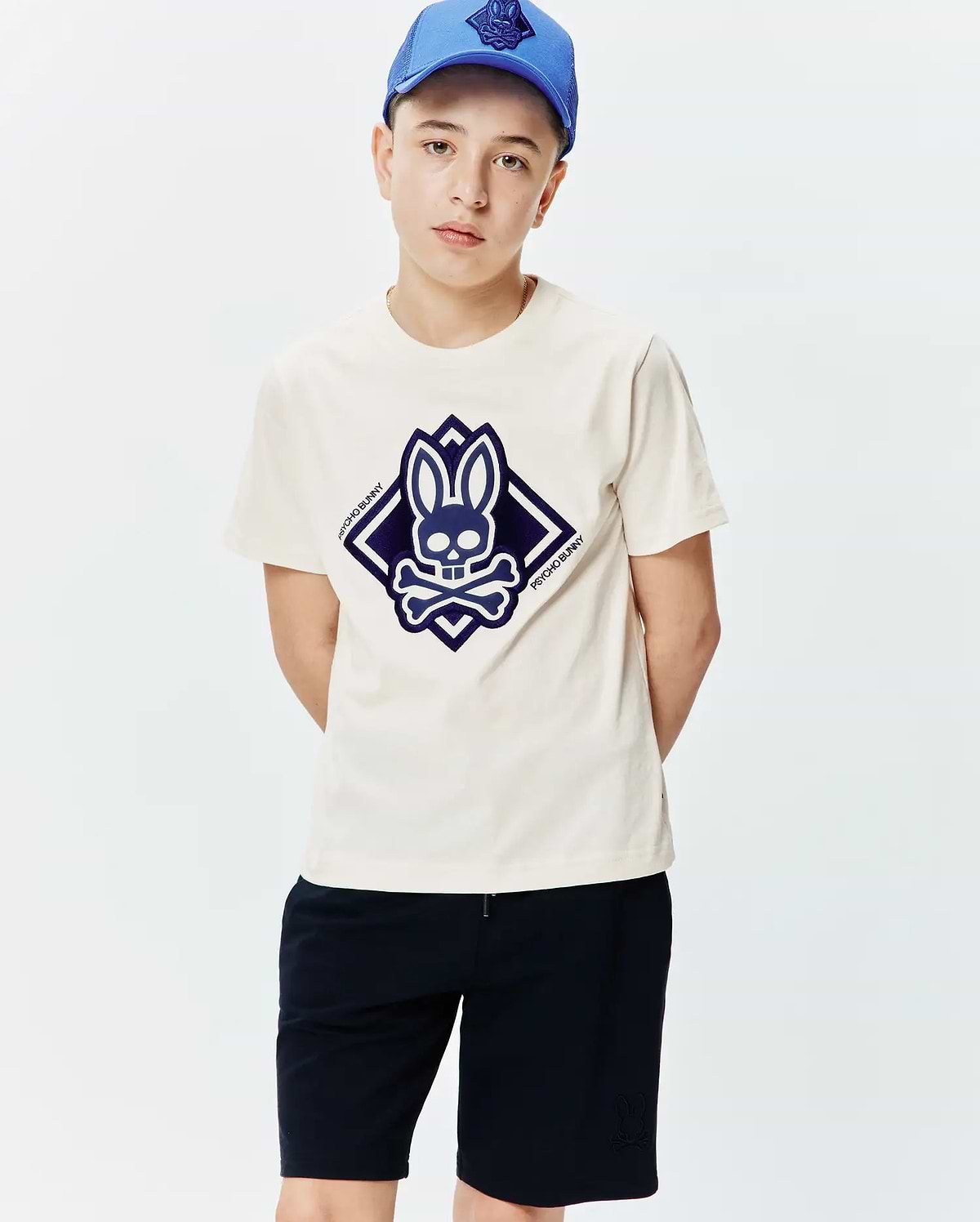 Kids Tobias Graphic Tee, a classic fit cream jersey t-shirt with a large central dark blue Psycho Bunny skull and crossbones logo graphic in a diamond shape.