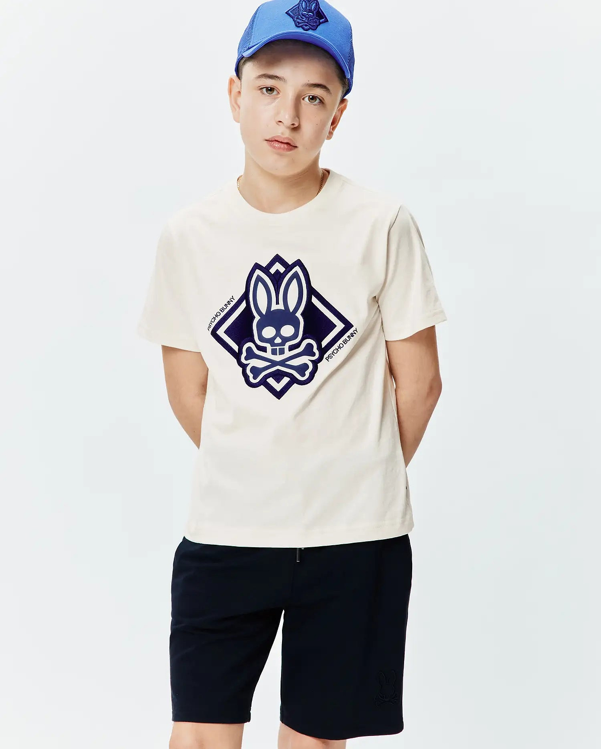 Kids Tobias Graphic Tee, a classic fit cream jersey t-shirt with a large central dark blue Psycho Bunny skull and crossbones logo graphic in a diamond shape.