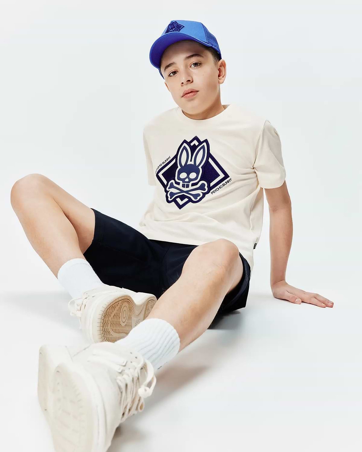 Kids Tobias Graphic Tee in cream jersey with a large navy blue Psycho Bunny skull logo on the chest, paired with navy shorts and a royal blue mesh baseball cap.