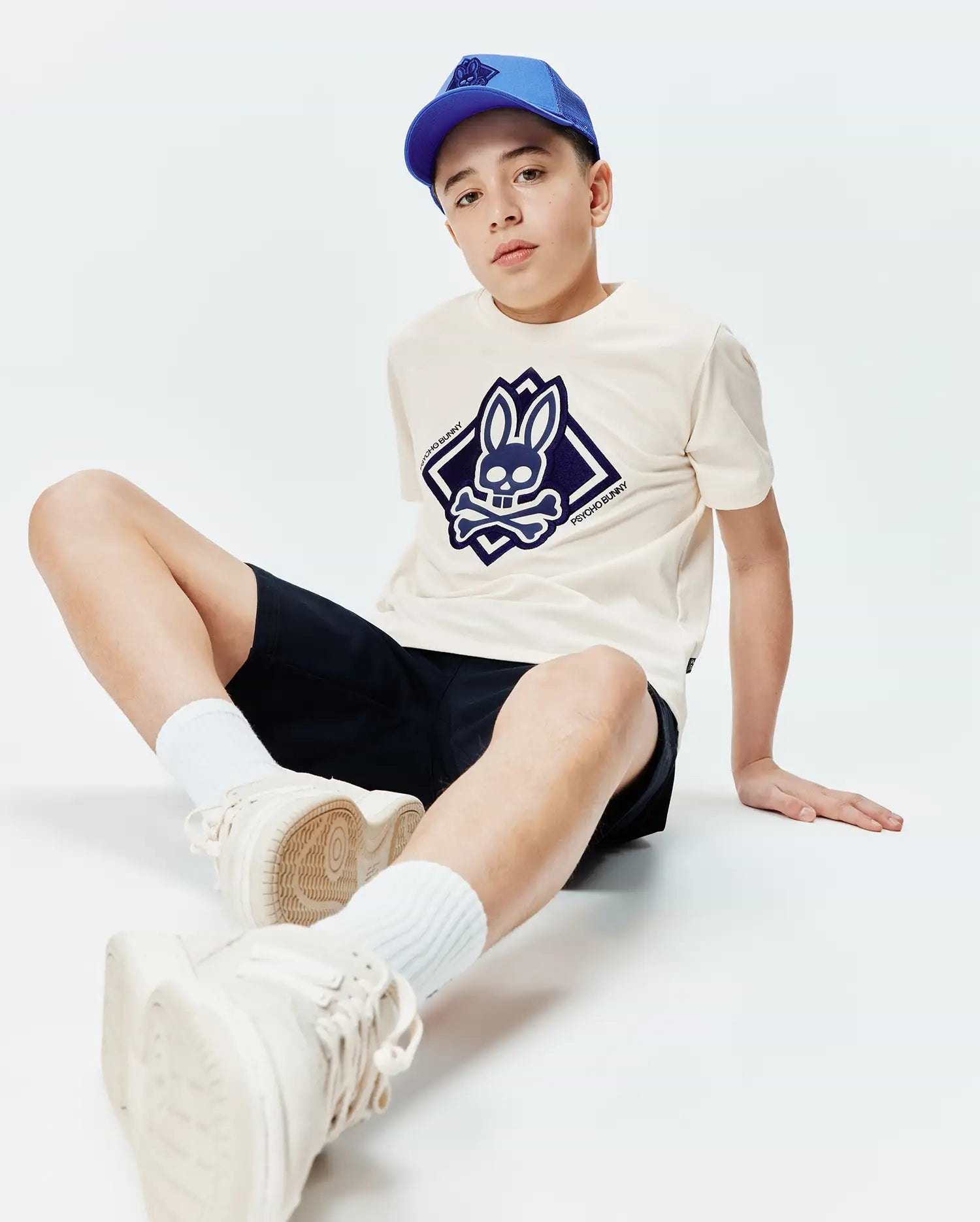 Kids Tobias Graphic Tee in cream jersey with a large navy blue Psycho Bunny skull logo on the chest, paired with navy shorts and a royal blue mesh baseball cap.