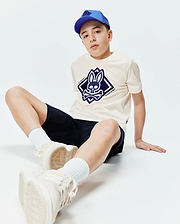 Kids Tobias Graphic Tee in cream jersey with a large navy blue Psycho Bunny skull logo on the chest, paired with navy shorts and a royal blue mesh baseball cap.