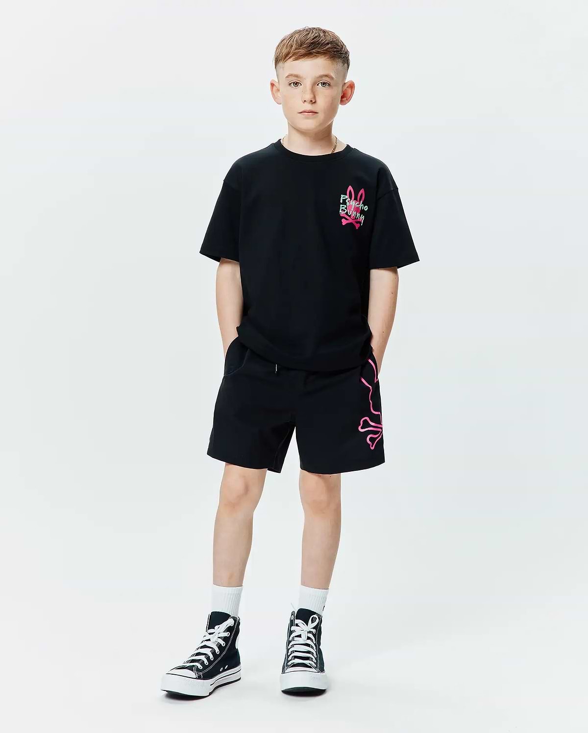 Kids Duncan Relaxed Fit Tee in black jersey with a small neon pink embroidered Psycho Bunny logo on the chest, paired with black relaxed fit shorts featuring a large neon pink outline bunny skull graphic on the left leg.