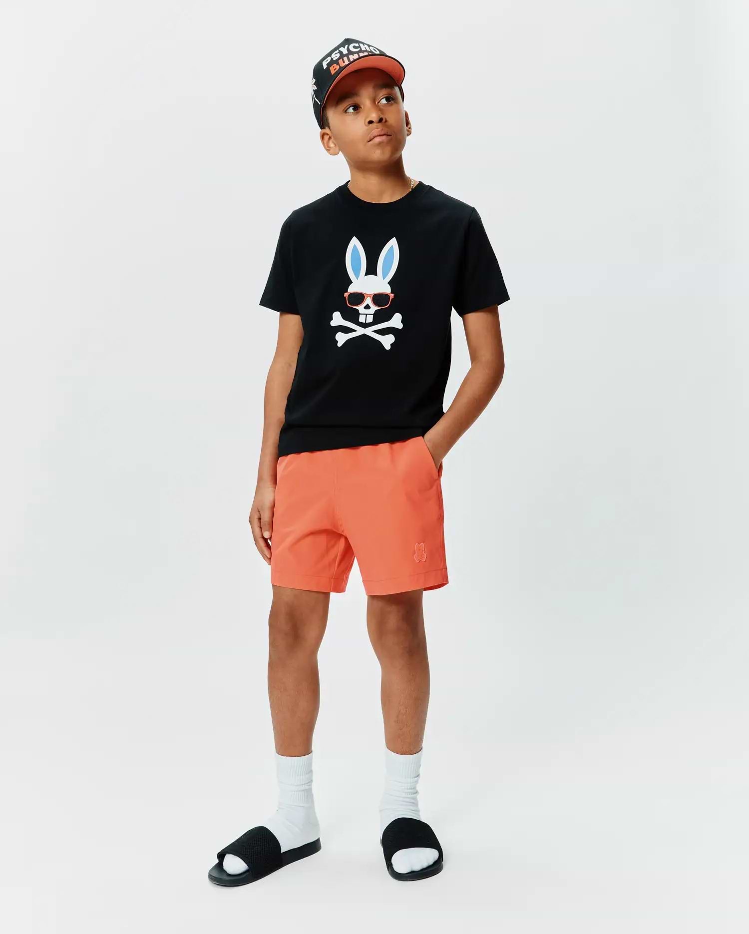 Kids Sunny Graphic Tee, a black classic fit jersey t-shirt with a large Psycho Bunny skull graphic featuring blue ears and red sunglasses on the chest, paired with orange shorts with a small embroidered bunny logo.