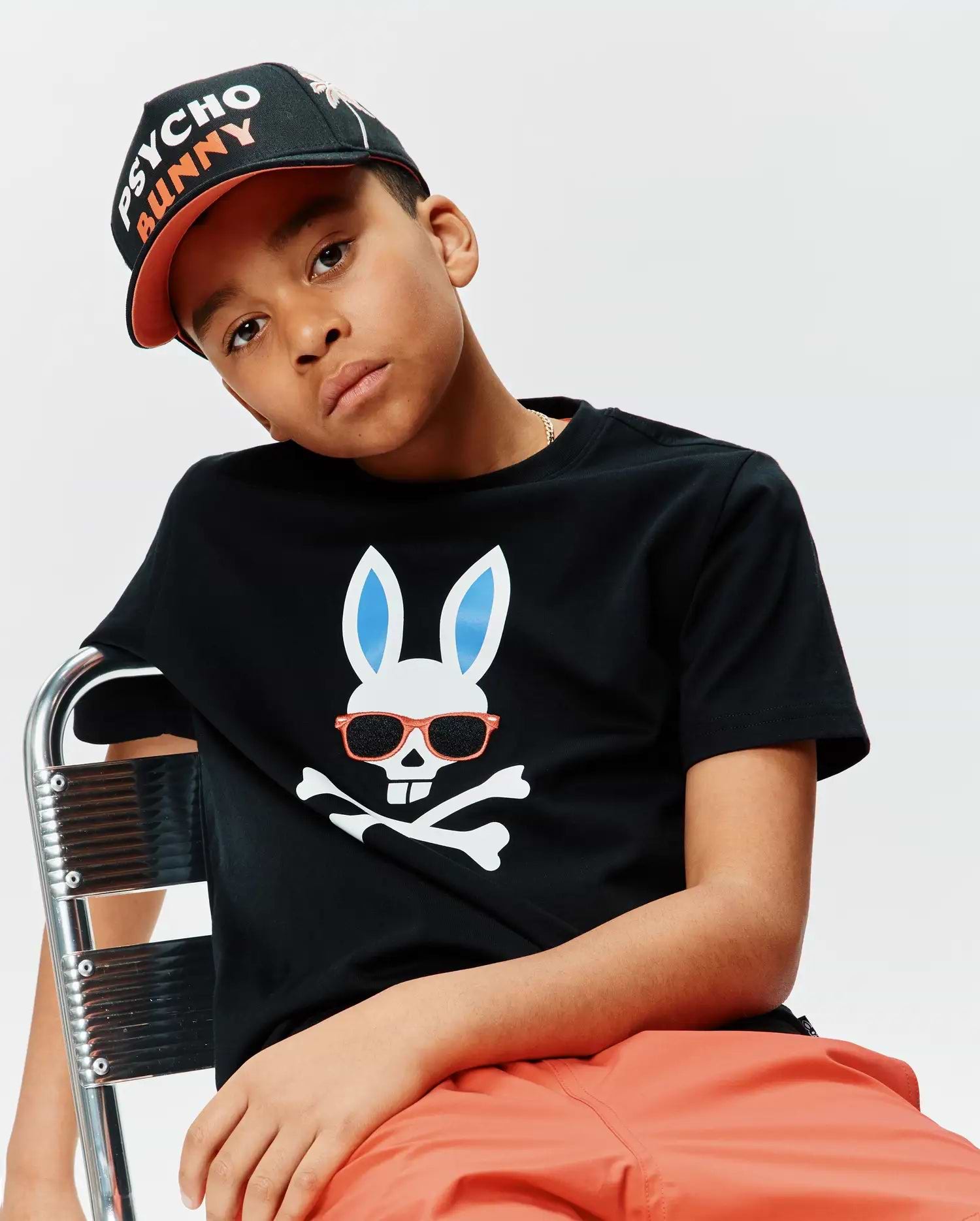 Kids Sunny Graphic Tee, a black classic fit jersey t-shirt featuring a large white Psycho Bunny skull graphic with light blue ears and orange sunglasses over crossbones, paired with a black and orange Psycho Bunny embroidered baseball cap.