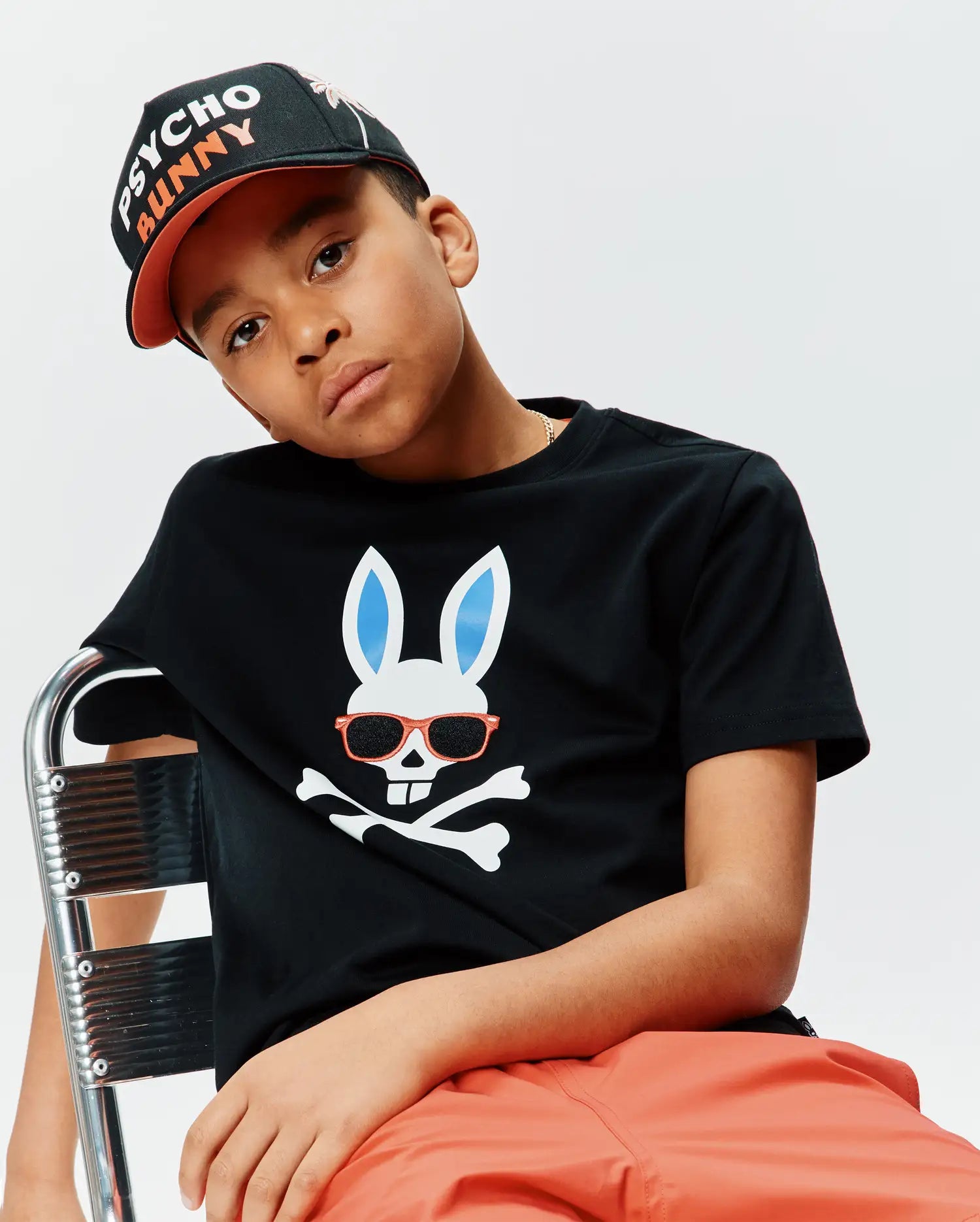 Kids Sunny Graphic Tee, a black classic fit jersey t-shirt featuring a large white Psycho Bunny skull graphic with light blue ears and orange sunglasses over crossbones, paired with a black and orange Psycho Bunny embroidered baseball cap.