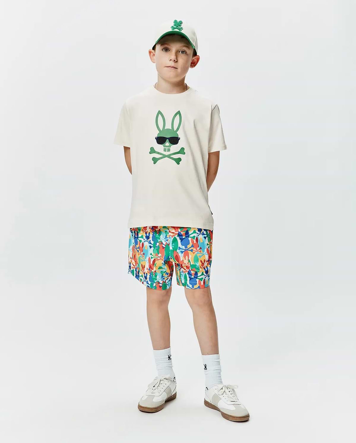Ecru jersey Kids Sunny Graphic Tee featuring a large green Psycho Bunny skull graphic with sunglasses, complemented by multicolor all-over bunny print shorts and a white embroidered logo cap.