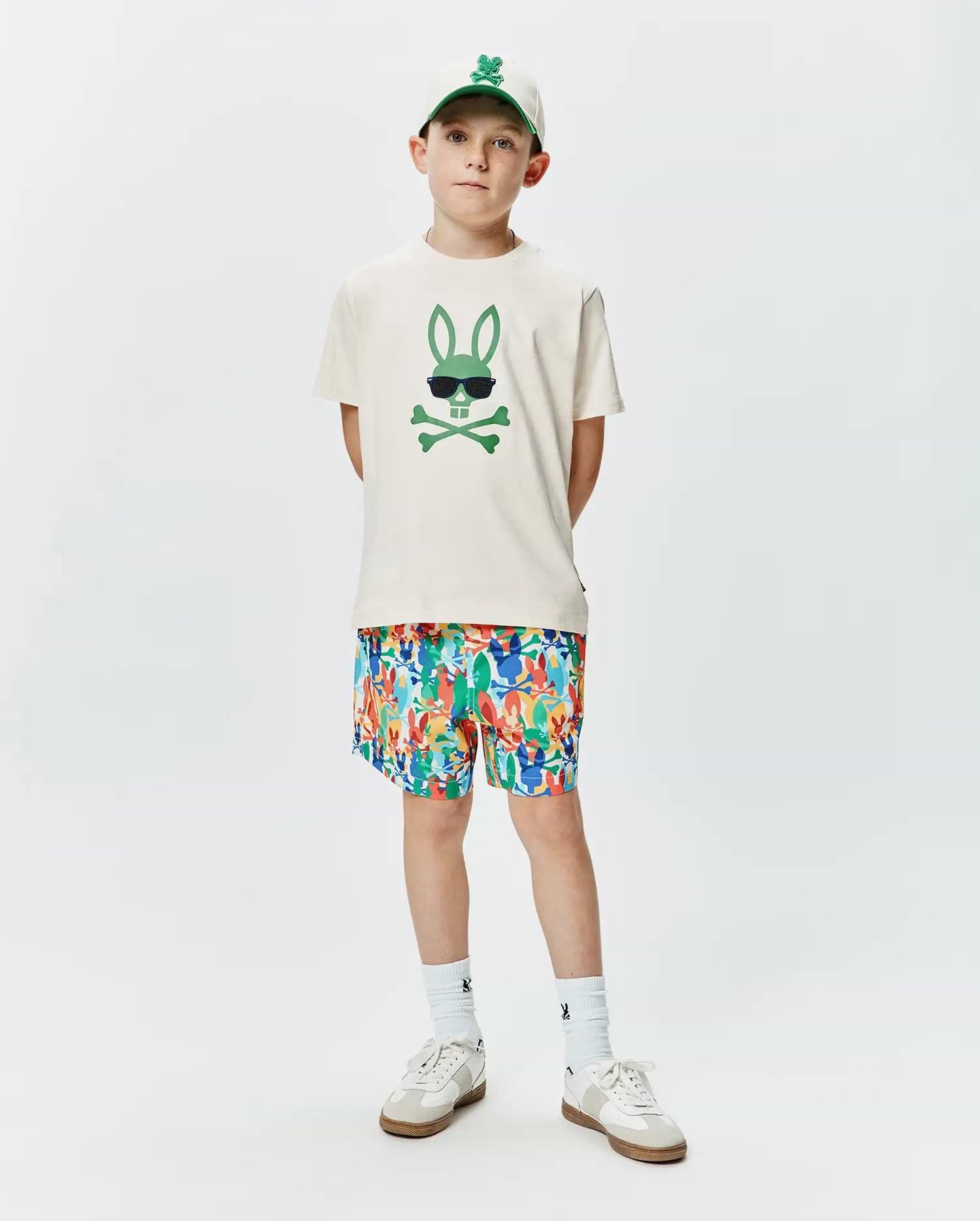 Ecru jersey Kids Sunny Graphic Tee featuring a large green Psycho Bunny skull graphic with sunglasses, complemented by multicolor all-over bunny print shorts and a white embroidered logo cap.