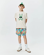 Ecru jersey Kids Sunny Graphic Tee featuring a large green Psycho Bunny skull graphic with sunglasses, complemented by multicolor all-over bunny print shorts and a white embroidered logo cap.