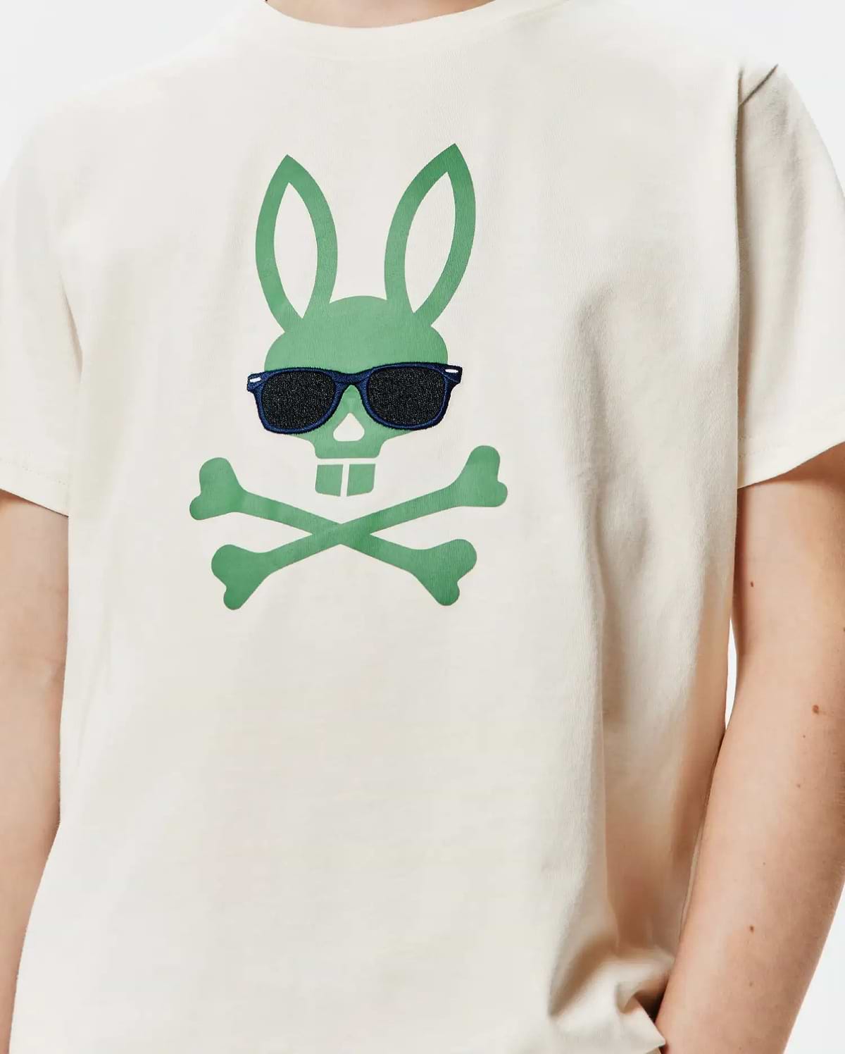 Kids Sunny Graphic Tee, a cream classic-fit jersey t-shirt featuring a large central green Psycho Bunny skull and crossbones logo wearing embroidered dark blue sunglasses with black lenses.