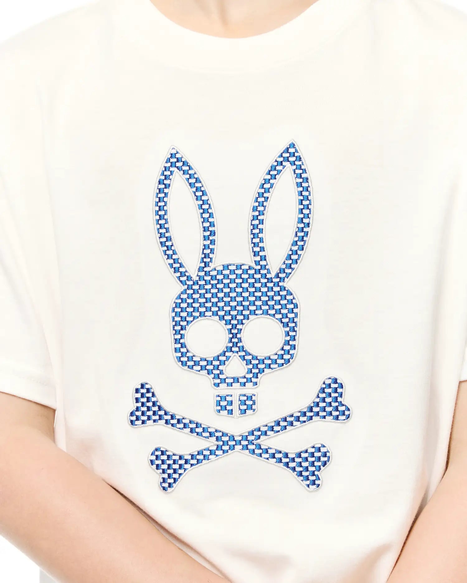 Kids Stan Graphic Tee, an off-white jersey t-shirt featuring a large, central embroidered Psycho Bunny skull and crossbones logo with a blue and white checkered pattern.