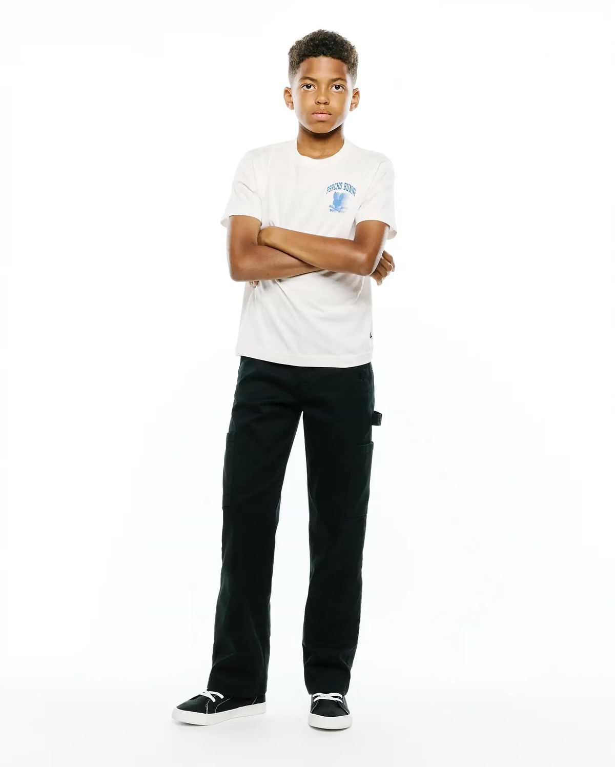 Kids Nathaniel Back Graphic Tee in off-white jersey, classic fit, featuring a light blue Psycho Bunny skull logo and text on the left chest, paired with dark green straight-leg pants.