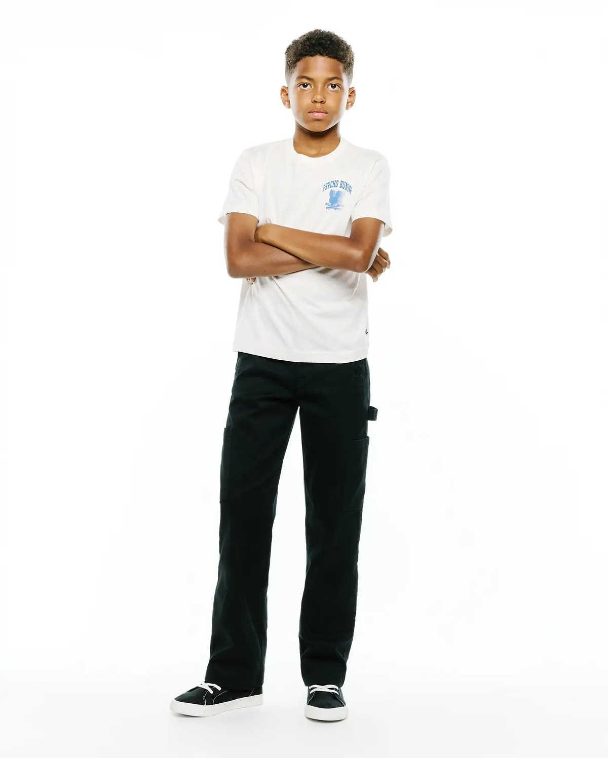 Kids Nathaniel Back Graphic Tee in off-white jersey, classic fit, featuring a light blue Psycho Bunny skull logo and text on the left chest, paired with dark green straight-leg pants.