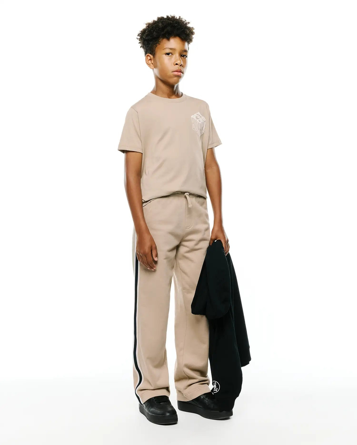 Beige jersey KIDS CONAN BACK GRAPHIC TEE with a white dripping Psycho Bunny chest graphic, matching relaxed fleece sweatpants with black and white side stripes, and a dark hoodie with a sleeve logo.