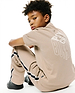 Kids Conan Back Graphic Tee, beige jersey with a large white 3D Psycho Bunny graphic on back, and relaxed beige track pants featuring black/white side stripes and an embroidered bunny skull.