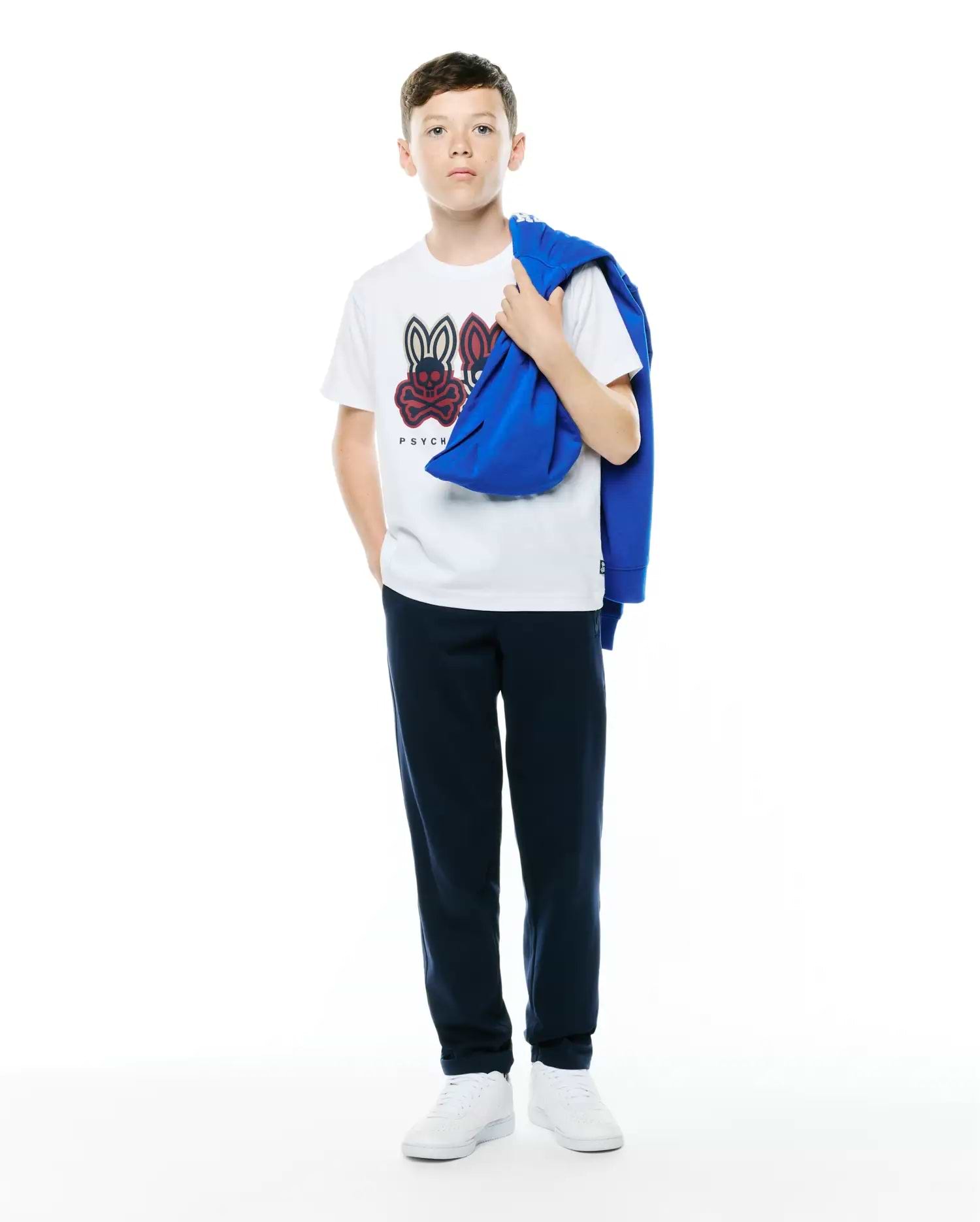 White Kids Ledger Graphic Tee featuring a large central red and black Psycho Bunny skull logo, styled with navy relaxed-fit sweatpants and a royal blue french terry hoodie.