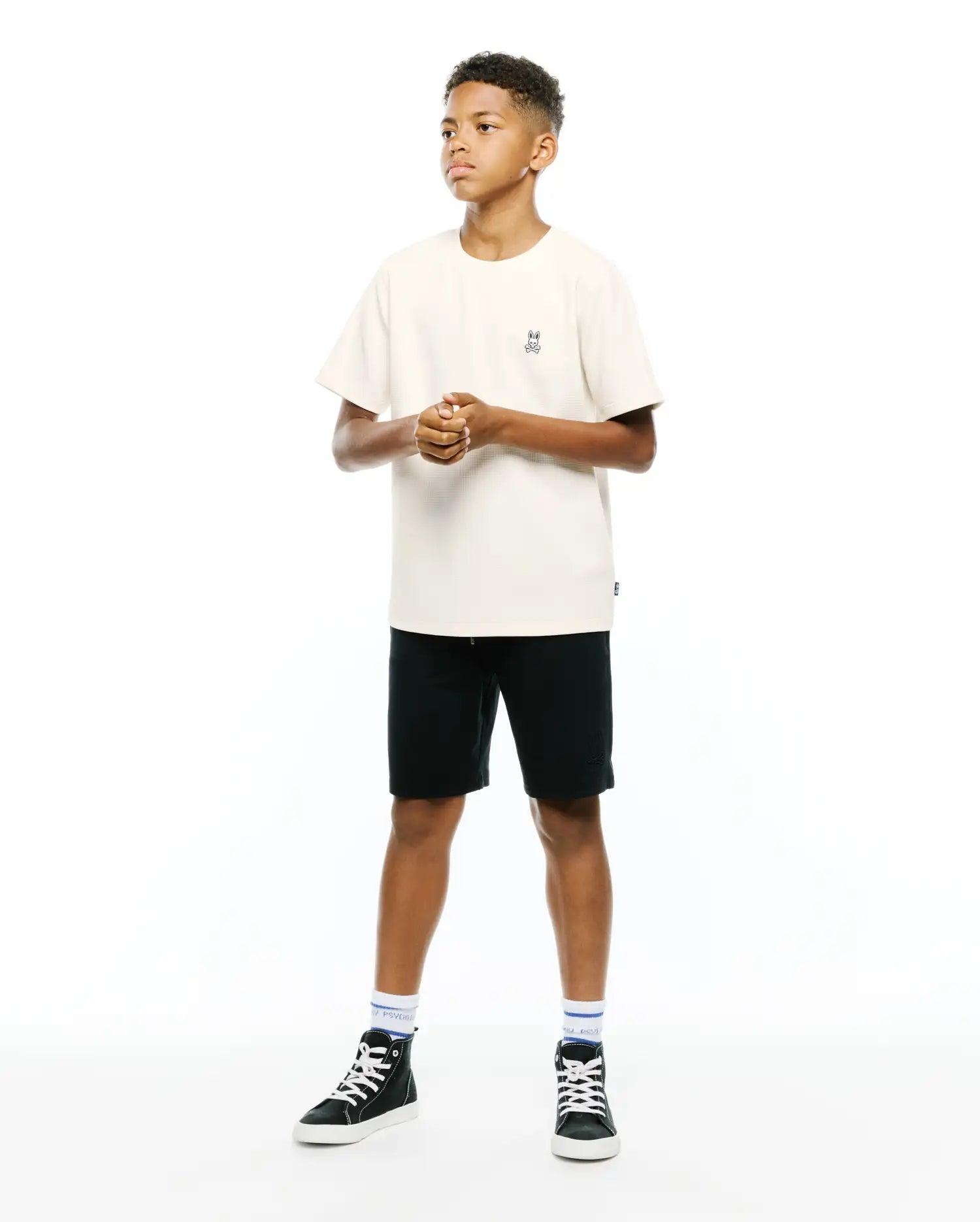 Kids Maxton Waffle Tee in cream, relaxed fit, waffle knit texture, with a small embroidered Psycho Bunny skull logo on the left chest. Black shorts feature a subtle embroidered logo.