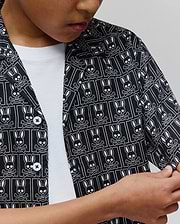 Kids Keon Tencel Shirt in black with an all-over white Psycho Bunny skull and crossbones logo pattern, featuring a relaxed fit, smooth Tencel fabric, and white buttons.