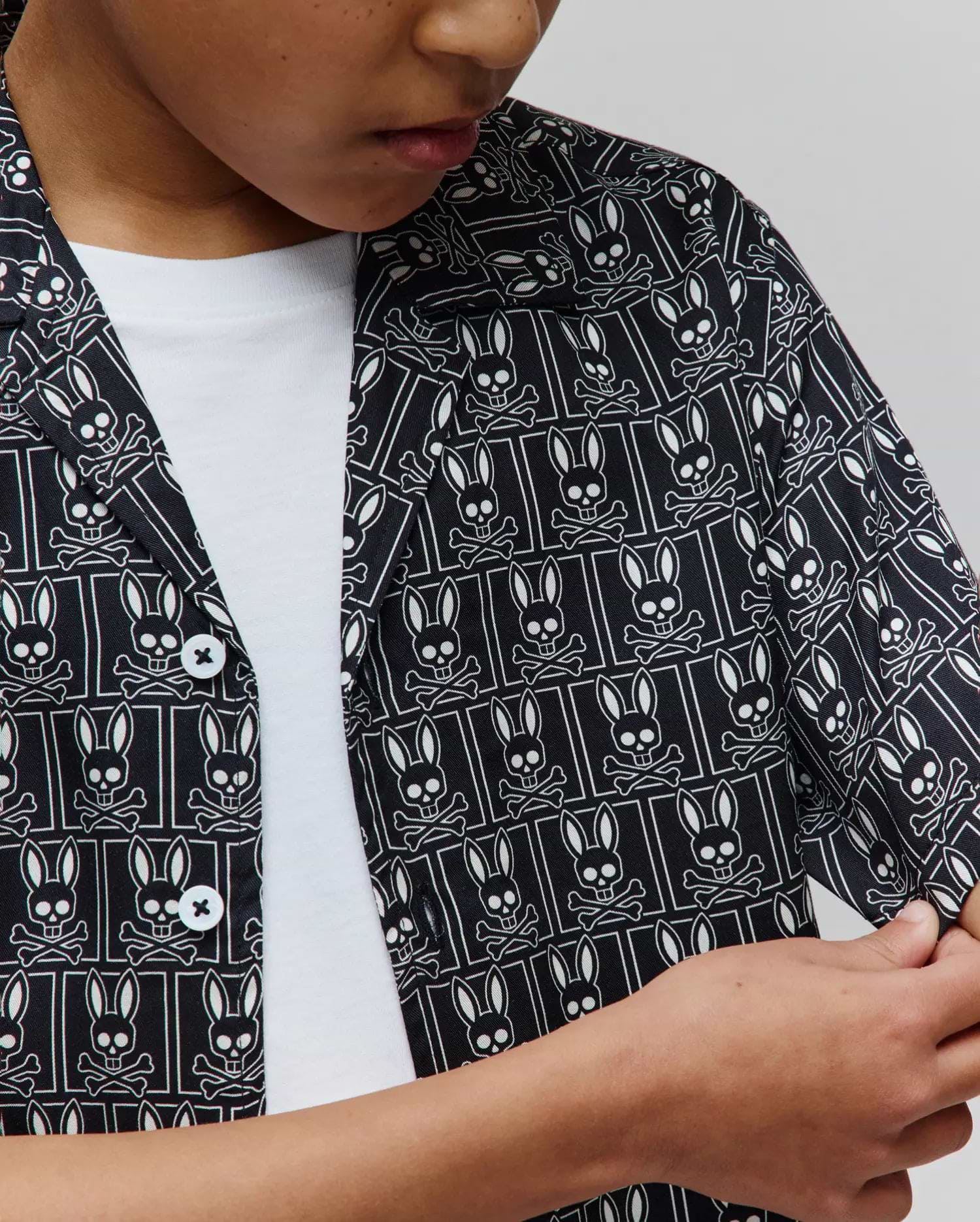 Kids Keon Tencel Shirt in black with an all-over white Psycho Bunny skull and crossbones logo pattern, featuring a relaxed fit, smooth Tencel fabric, and white buttons.