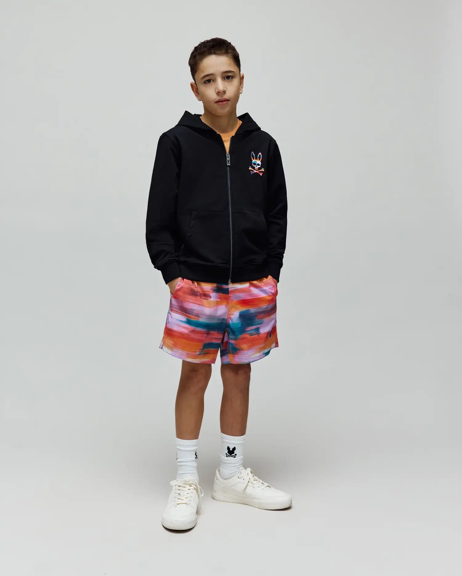 Kids Raphael French Terry Full Zip Hoodie in black with a colorful embroidered Psycho Bunny logo on the chest, paired with vibrant multi-color abstract print shorts and white socks featuring a small black bunny logo.
