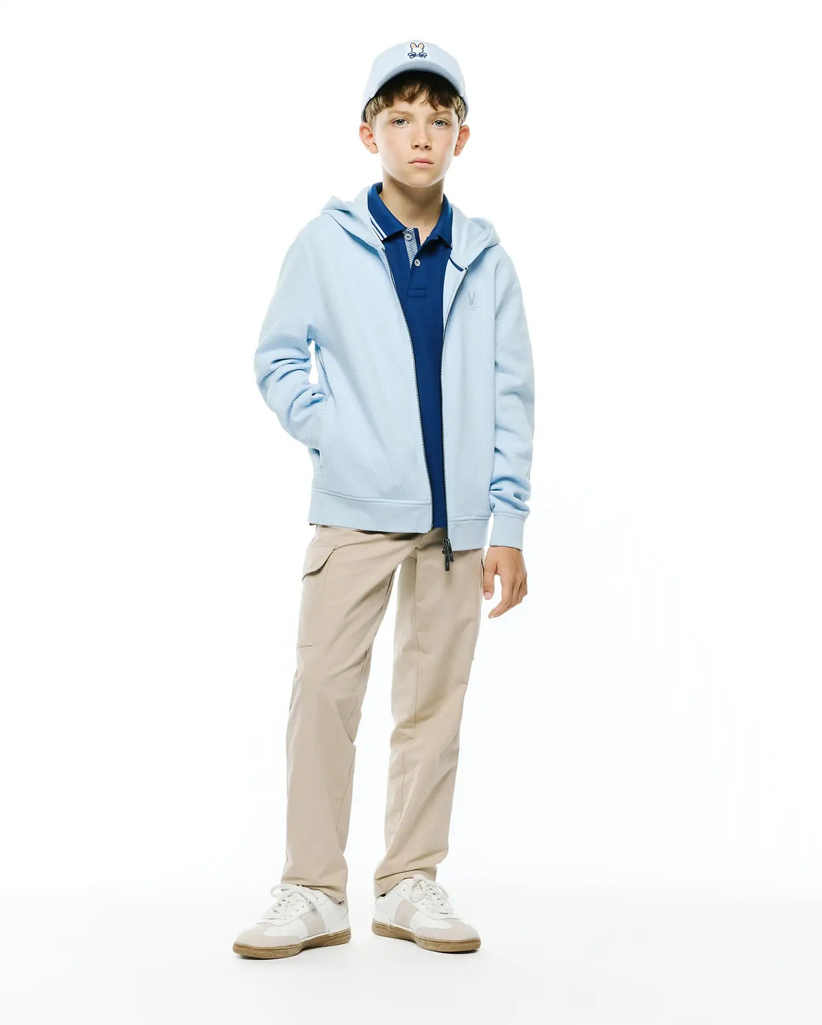 Kids Marc Textured Full Zip Hoodie in light blue, with textured fabric and an embroidered Psycho Bunny chest logo. Layered over a navy pique polo, beige cargo pants, and a light blue Psycho Bunny cap.