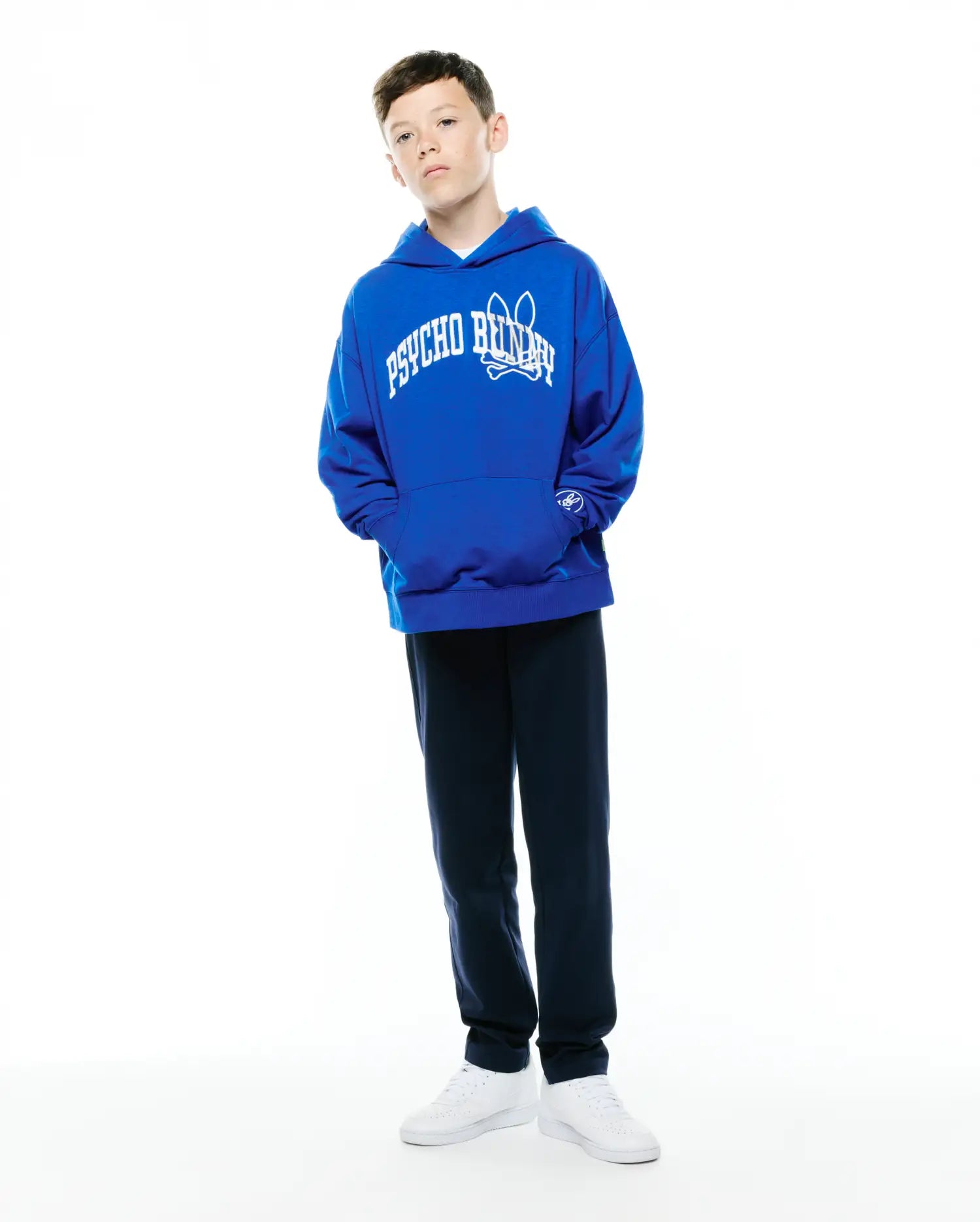 Kids Nathaniel Relaxed Hoodie, royal blue French terry, features a large white arched PSYCHO BUNNY text and bunny skull logo on the chest, plus a small embroidered bunny skull on the kangaroo pocket.